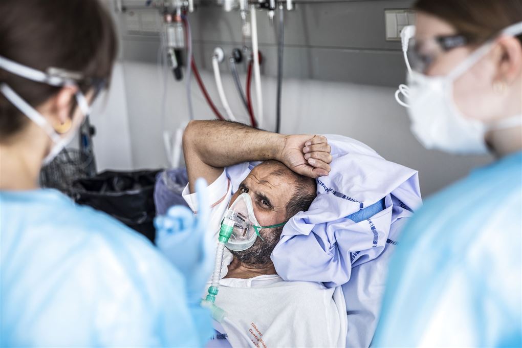 En patient ligger i hospitalsseng med iltmaske på mens to sygeplejersker med værnemidler ser til ham 