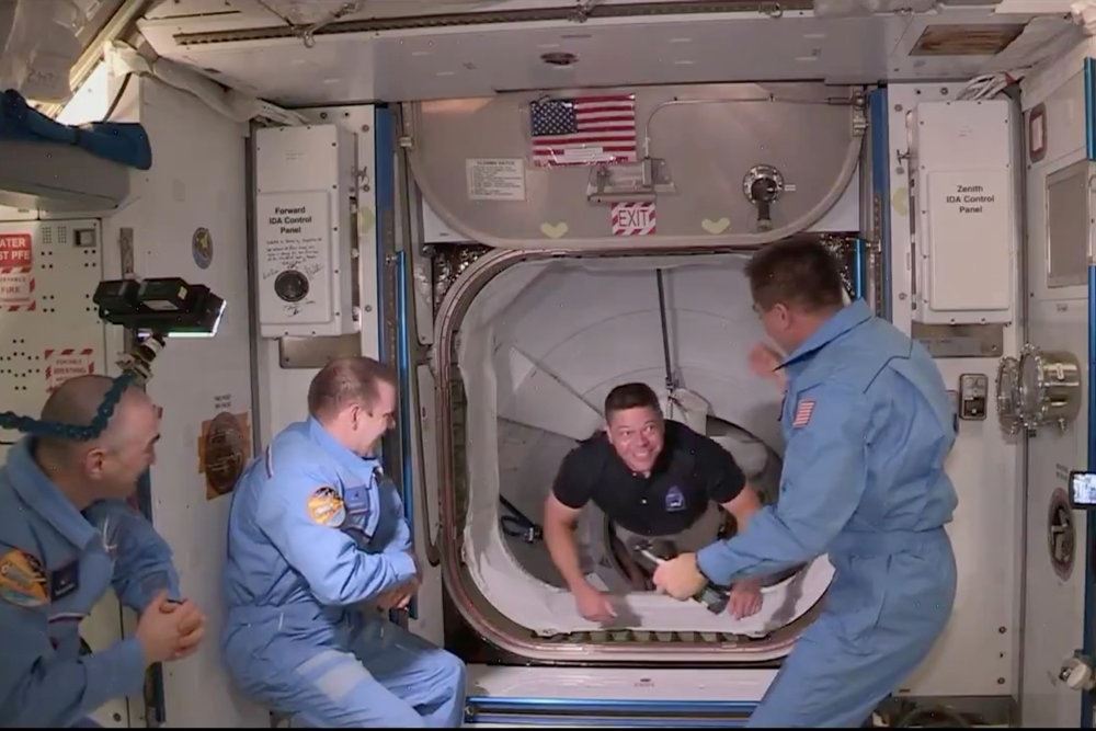 En astronaut kravler igennem slusen på Rumstationen ISS og bliver modtaget af tre andre astronauter