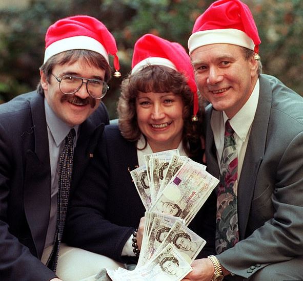 Tre personer med nissehuer griner bredt, mens de sidder med en sæk fyldt med pengesedler