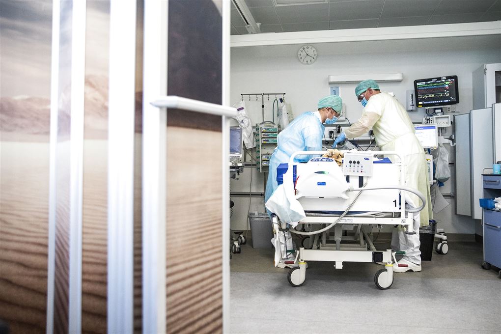 To læger eller sygeplejersker står bøjet over en hospitalsseng. De er iført beskyttelsestøj som kitler, handsker og hårnet.