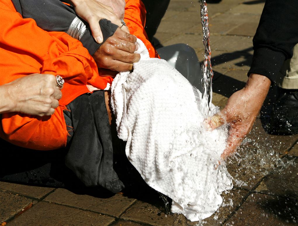 En person ligger på jorden med et hvidt klæde foran ansigtet og hænderne bundet med gaffatape. Der plasker vand ned i ansigtet på vedkommende, som også bliver holdt nede på billedet. 