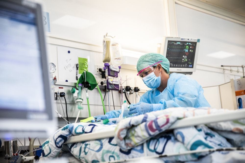 En sygeplejerske med mundbind, hætte og handsker på står ved en patient på intensivafdelingen.