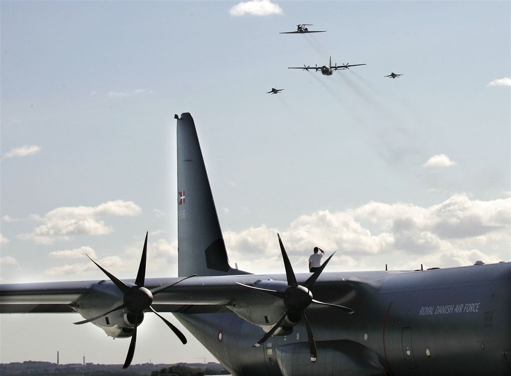 Et stort gråt Herkulesfly holder i en lufthavn. I baggrunden ses flere andre fly i luften. 