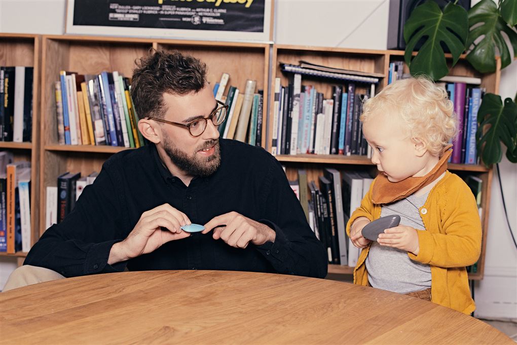 En far med skæg og briller sidder sammen med en lille pige med krøller og gul bluse med bøger i en reol bagved