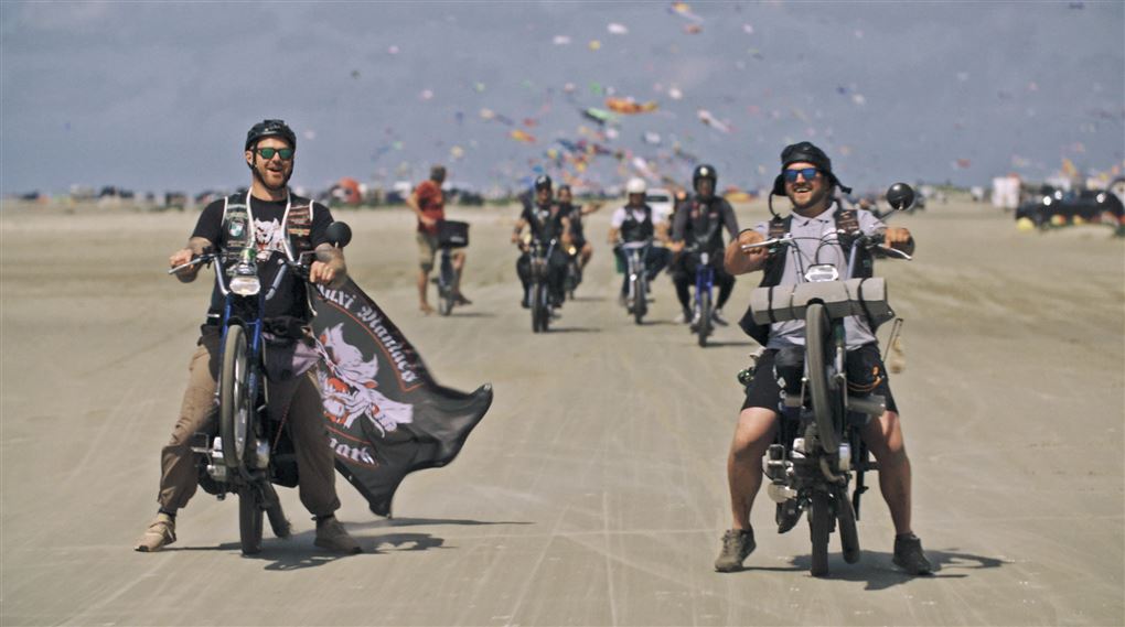 En flok motorcykelister kører på en bred grusvej. Der er konfetti i luften omkring dem.