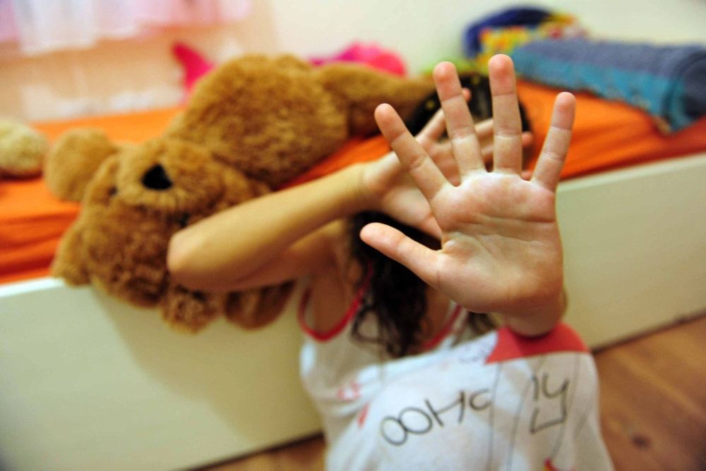 En pige holder sine hænder foran sit ansigt. Hun sidder på gulvet foran en seng, hvor der ligger en tøjbamse.