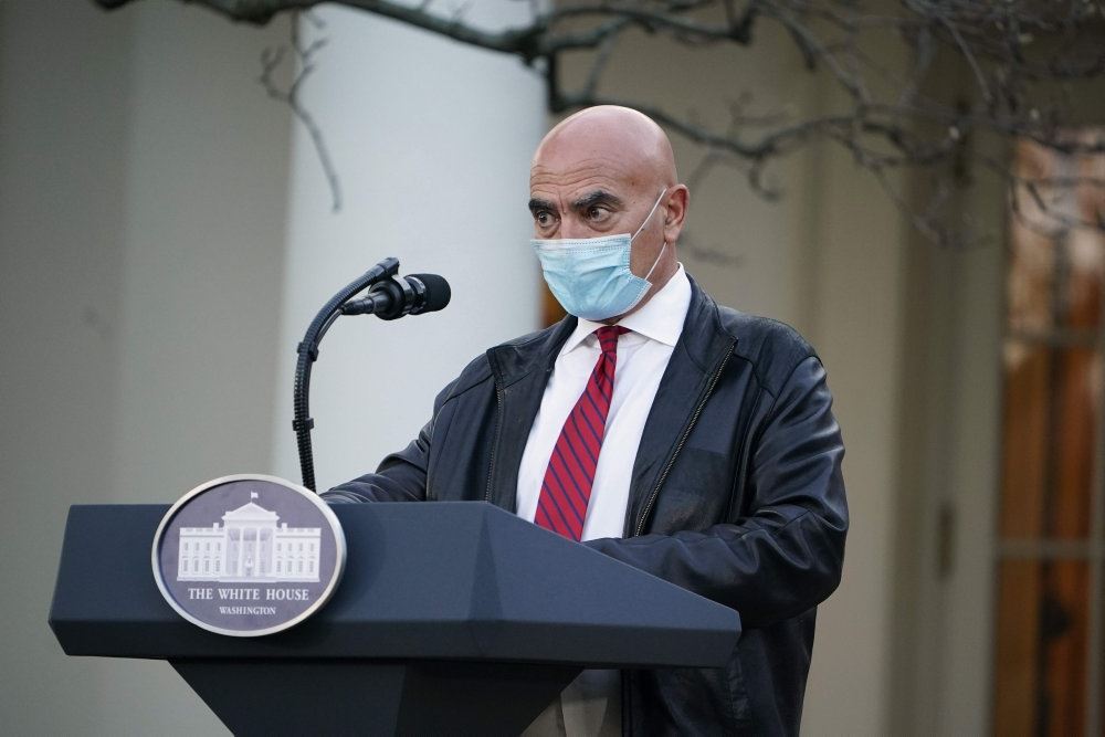 En mand med maske i jakkesæt på en talerstol