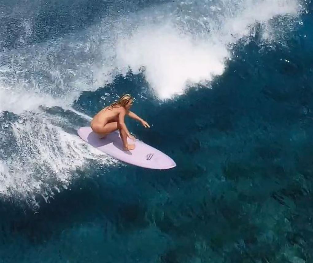En nøgen kvinde står på surfbræt på en bølge