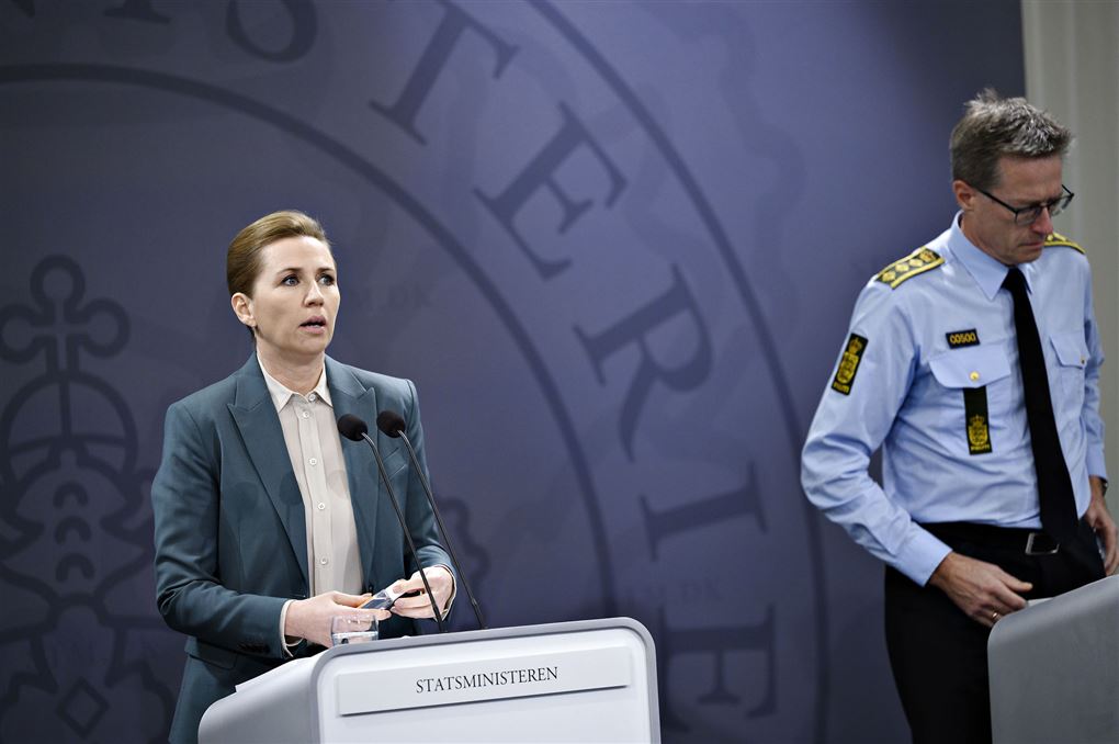 Mette Frederiksen ved dagens coroa-pressemøde i baggrunden ses politiet.