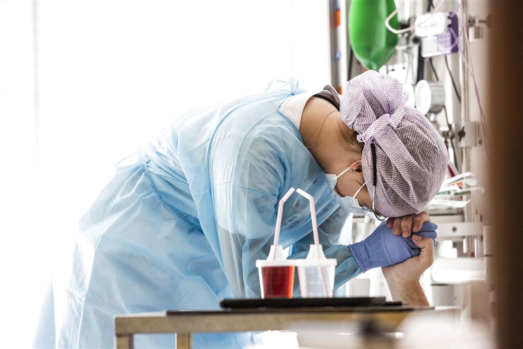 En sygeplejerske med engangskittel, maske, hårnet og gummehandsker holder en patiens hånd på sin pande. 