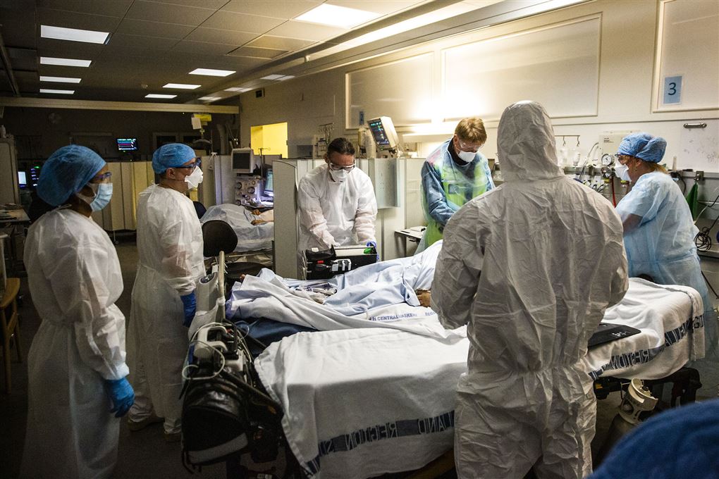 En flok læger og sygeplejersker i fuld værnemiddel outfit står rundt om en hospitalsseng på en corona-afdeling.
