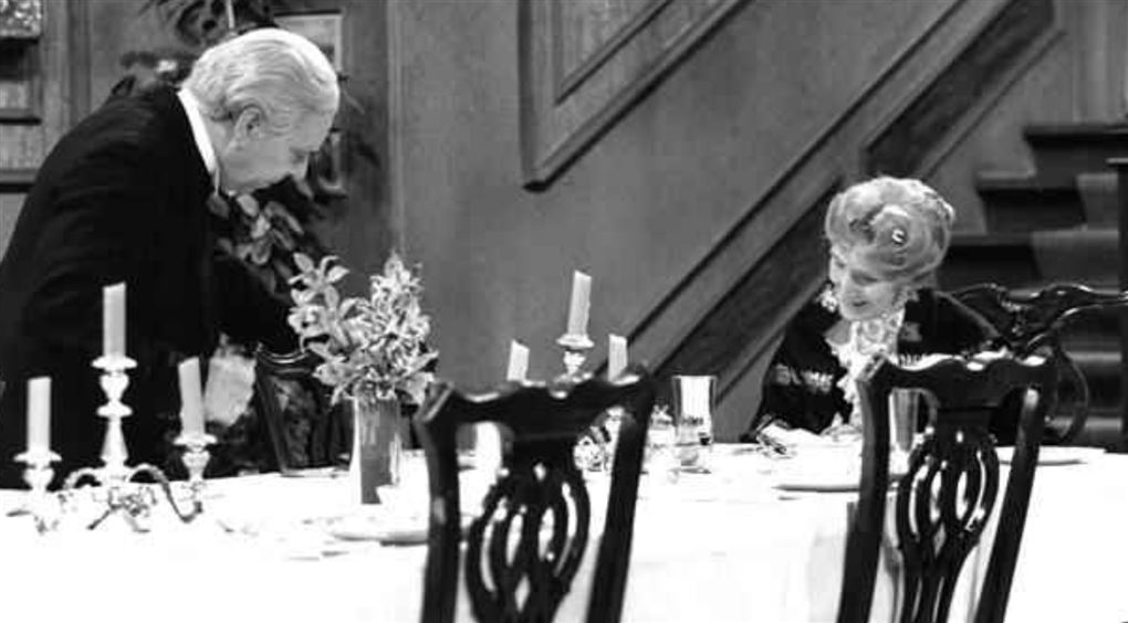 En ældre tjener hælder op for en ældre dame siddende ved et veldækket bord