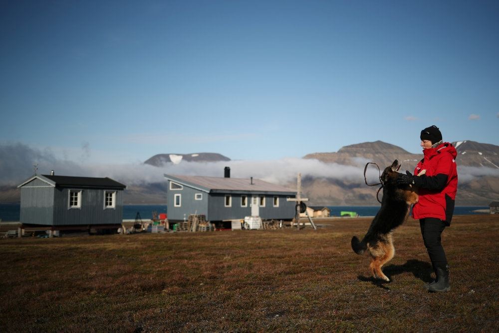 En mand med en hund foran en hytte med fjelde i baggrunden