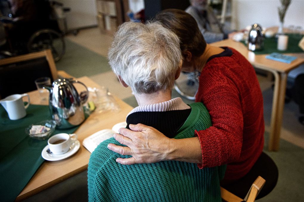 en gammel dame sidder med ryggen til mens en kvinde lægger armen om hendes ryg.
