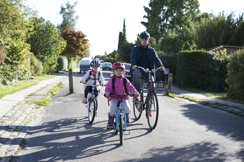 En far cykler med to små børn - alle bærer hjelm.